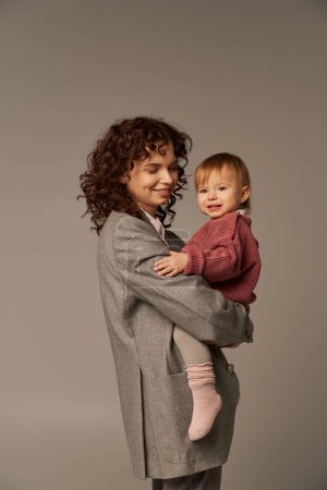 quality family time, parenting and career, cheerful businesswoman holding in arms her toddler daughter on grey background, work life harmony concept, working parent, loving motherhood