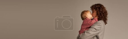 Photo for Parenting and career, curly businesswoman holding in arms and kissing her toddler daughter on grey background, work life harmony concept, loving motherhood, quality time, side view, banner - Royalty Free Image