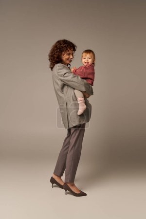 quality family time, parenting and career, joyful businesswoman holding in arms her toddler daughter on grey background, work life harmony concept, working parent, loving motherhood, full length 