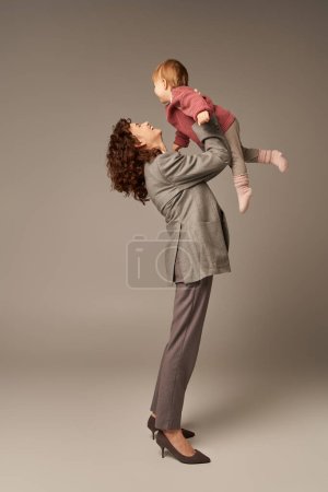 quality time, parenting and career, cheerful businesswoman lifting her toddler daughter on grey background, work life harmony concept, working parent, loving motherhood, full length 