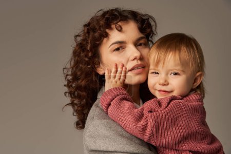 working mother, parenting and career, curly businesswoman embracing cute toddler daughter on grey background, work life harmony concept, loving motherhood, quality family time 