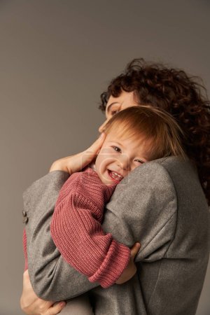 working mother, parenting and career, curly businesswoman embracing happy toddler daughter on grey background, work life harmony concept, loving motherhood, quality family time 