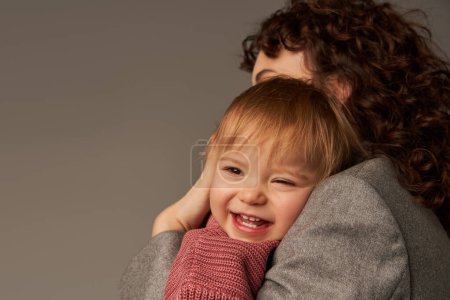 parenting and career, curly businesswoman embracing happy toddler daughter on grey background, work life harmony concept, loving motherhood, quality family time, working mother 