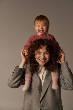 motherhood, work life balance, cheerful working mother with kid on shoulders, woman and happy child on grey background, work life harmony concept, loving motherhood, quality time 