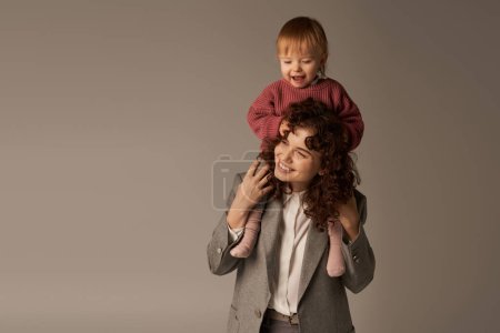 motherhood, kid on shoulders, parenting and career, curly businesswoman and happy daughter on grey background, work life harmony concept, loving motherhood, quality family time, work life harmony