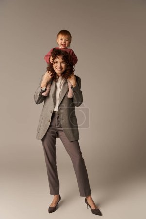 Photo for Kid on shoulders, loving motherhood, parenting and career, curly businesswoman and happy toddler daughter on grey background, work life harmony concept, loving motherhood, quality family time - Royalty Free Image