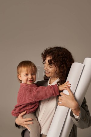 Aufbau einer erfolgreichen Karriere, Multitasking-Frau, berufliche Errungenschaften, Zeitmanagement, fröhliche Mutter, die ihre Tochter im Arm hält und gerollte Papierentwürfe auf grauem Hintergrund 