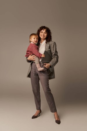 professional aspirations, modern parenting, work life harmony, happy working mother holding laptop and daughter on grey background, time management, self-confidence, full length 