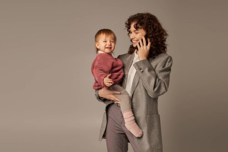 Photo for Modern parenting, building successful career, empowered woman, cheerful mother talking on smartphone and holding in arms toddler daughter on grey background, motherhood, businesswoman - Royalty Free Image