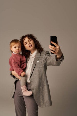 modern parenting, building successful career, empowered woman, curly mother taking selfie on smartphone and holding in arms toddler daughter on grey background, motherhood 