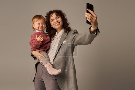 modern parenting, work life balance, empowered woman, happy mother taking selfie on smartphone and holding in arms toddler daughter on grey background, motherhood 