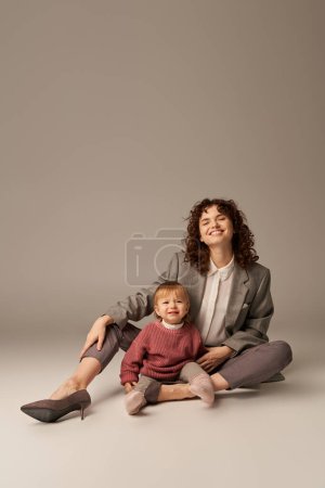 career and family, balancing between work and life, happy woman with curly hair hugging toddler daughter on grey background, quality time, modern parenting, motherhood, full length  
