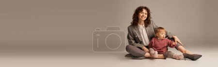 career and family, balancing between work and life, cheerful mother with curly hair hugging toddler daughter on grey background, quality time, modern parenting, businesswoman, full length, banner 