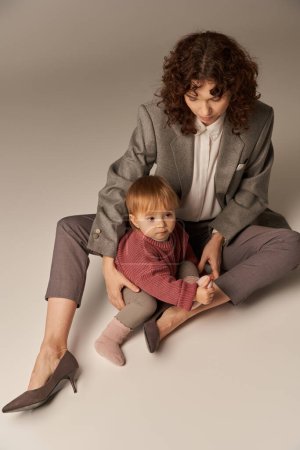 Photo for Career and family, balancing between work and life, cheerful mother with curly hair hugging toddler daughter on grey background, quality time, modern parenting, businesswoman, high angle view - Royalty Free Image