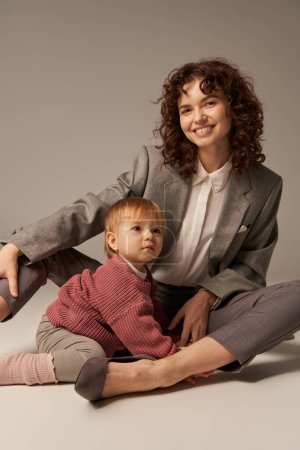 career and family, balanced lifestyle, cheerful mother with curly hair hugging toddler daughter on grey background, quality time, modern parenting, businesswoman, loving motherhood  