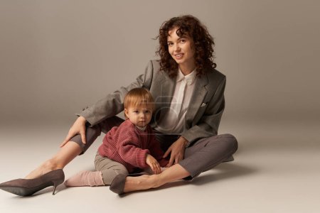 parenting and family, balanced lifestyle, cheerful mother with curly hair hugging toddler daughter on grey background, quality time, modern woman, businesswoman, loving motherhood  