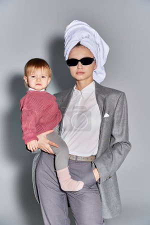 Multitasking, Frau mit Sonnenbrille, Kleinkind im Arm und mit Handtuch auf dem Kopf, Balanceakt zwischen Arbeit und Leben, ermächtigte Frau in formeller Kleidung auf grauem Hintergrund 