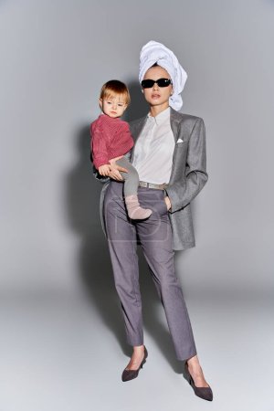 multitasking, woman in sunglasses holding in arms toddler daughter and standing with towel on head, balancing between work and life, empowered woman in formal wear on grey background, full length 
