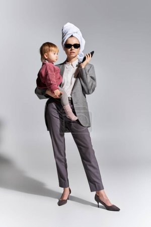 Multitasking, Frau mit Sonnenbrille, Tochter im Arm und mit Handtuch auf dem Kopf, balancierender Lebensstil, Geschäftsfrau in formeller Kleidung, Smartphone auf grauem Hintergrund, in voller Länge