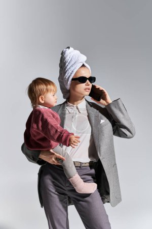 telefon, wielozadaniowość, kobieta w okularach przeciwsłonecznych trzymająca w ramionach córkę i stojąca z ręcznikiem na głowie, równoważąca styl życia, businesswoman w formalnym ubraniu rozmawiająca na smartfonie na szarym tle 