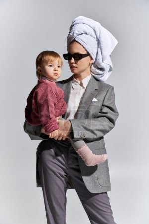 kobieta w okularach przeciwsłonecznych trzymająca w ramionach córeczkę malucha i stojąca z ręcznikiem na głowie, wielozadaniowość, równowaga między pracą a życiem, uzdolniona kobieta w formalnym ubraniu na szarym tle 