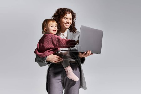 Zeitmanagement, berufstätige Mutter mit Laptop und Kleinkind im Arm vor grauem Hintergrund, Vereinbarkeit von Beruf und Familie, moderne Elternschaft, beruflicher Erfolg, fröhliche Geschäftsfrau 