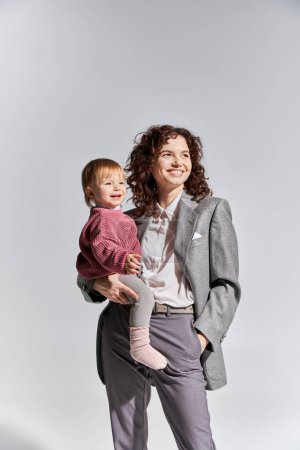 career and family, happy working mother holding toddler daughter in arms and standing with hand in pocket on grey background, work life balance, modern parenting, professional success, businesswoman 