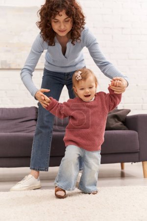first steps, quality time, bonding, balancing work and life, family relationships, working mother holding hands with toddler daughter, togetherness, cozy living room, denim jeans, casual attire