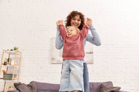 engaging with child, balancing work and life, family bonding, happy and curly mother lifting toddler daughter, togetherness, cozy living room, quality time, denim jeans, loving motherhood 