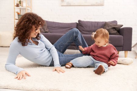 Work-Life-Balance, glückliche und lockige Frau auf Teppich sitzend mit aufgeregter Kleinkind-Tochter im gemütlichen Wohnzimmer, hochwertige Familienzeit, lässige Kleidung, Bindung zwischen Mutter und Kind 