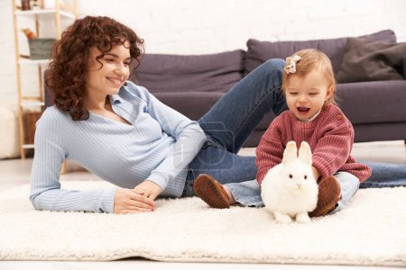 engageant avec enfant, femme heureuse et bouclée assise sur le tapis avec la fille en bas âge dans le salon confortable, jouer avec le lapin, temps de famille de qualité, tenue décontractée, liaison entre la mère et l'enfant 