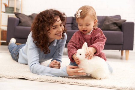 engaging with kid, cheerful and curly woman lying on carpet with toddler daughter in cozy living room, playing with rabbit, quality family time, casual attire, bonding between mother and child 