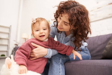 engaging with kid, happy mother hugging toddler daughter in cozy living room, playing with rabbit, quality time, casual attire, bonding between mother and child, stuffed animal 