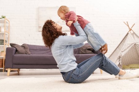 quality family time, curly woman lifting toddler daughter and sitting on carpet in cozy living room, work life balance, denim clothes, casual attire, family relationships, modern parenting 