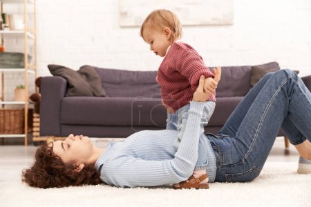 modern parenting mother lying on carpet and holding hands while supporting toddler daughter in cozy living room, work life balance, denim clothes, casual attire, engaging with kid, quality time 