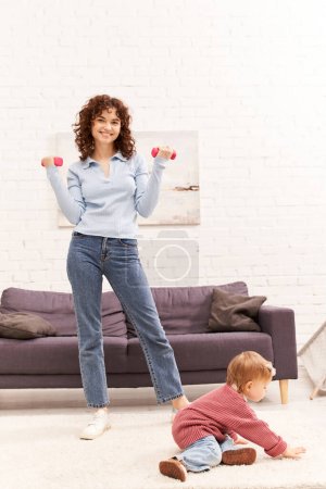 gestion du temps, mère au travail, mode de vie équilibré, femme bouclée faisant de l'exercice avec des haltères près de la petite fille dans le salon confortable, entraînement à la maison, sport, maman occupée, activité physique 