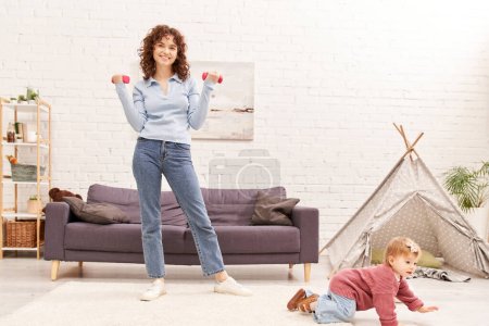 zarządzanie czasem, pracująca matka, zrównoważony styl życia, szczęśliwa kobieta ćwicząca z hantlami w pobliżu córki malucha w przytulnym salonie, trening domowy, sport, zajęta mama, aktywność fizyczna, wnętrze 