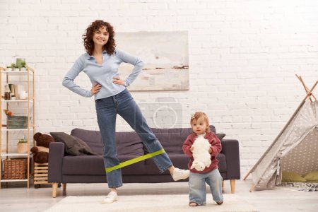 Photo for Busy mom, modern parenting, working mother, balanced lifestyle, happy woman exercising with resistance band near toddler daughter with soft toy in cozy living room, home workout, sport - Royalty Free Image