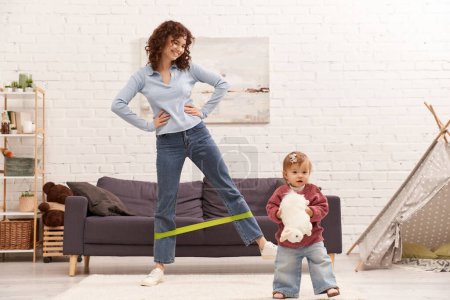 busy mom, home workout, sport, working mother, balanced lifestyle, happy woman exercising with resistance band near toddler daughter with soft toy in cozy living room, modern parenting 
