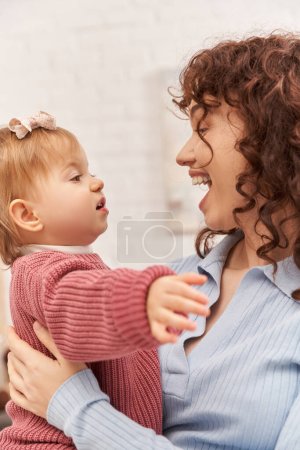 balanced lifestyle, working mother with curly hair holding in arms baby girl, amazed woman and her toddler daughter, modern parenting, work life harmony, bonding and engaging with kid, happiness 