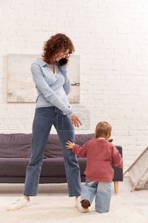 pełna długość malucha dziecko robi jej pierwsze kroki w pobliżu szczęśliwej matki, przytulny salon, angażując się z dzieckiem, dżinsy jeansowe, casual strój, czas rodzinny, nowoczesne rodzicielstwo, równowaga życia zawodowego 