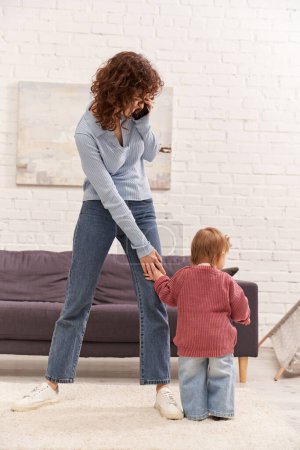 Photo for Full length of toddler baby doing her first steps near curly mother, engaging with kid, denim jeans, casual attire, family time, modern parenting, work life balance, cozy living room - Royalty Free Image