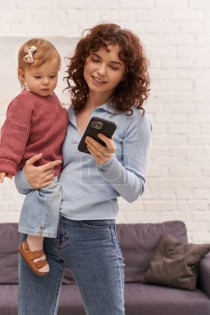 curly young woman holding in arms baby girl and using smartphone, balancing between work and life, modern parenting, relationships, mother daughter bond, remote work, career and family 