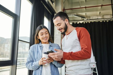 happy bearded man showing mobile phone to attractive asian woman holding coffee to go in office, interracial business colleagues, young entrepreneurs, startup project collaboration