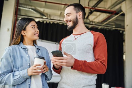 joyful asian woman with takeaway drink and bearded man with mobile phone smiling at each other in modern office, stylish casual clothes, successful partnership in business startup