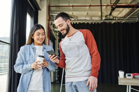 glad and bearded man networking on smartphone near smiling asian woman with takeaway drink, youthful and creative multiethnic business colleagues in modern office space