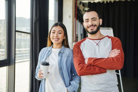 pleased asian woman with coffee to go in paper cup and bearded man with folded arms smiling at camera in modern office, young interracial entrepreneurs collaborating in startup 