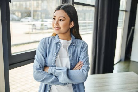 zufriedene asiatische Frau mit brünetten Haaren, die mit verschränkten Armen am Fenster in modernen Büroräumen steht, blaues Jeanshemd, glückliches Lächeln, junge und erfolgreiche Unternehmerin