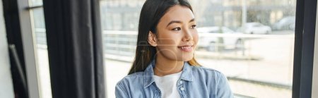 Photo for Attractive and joyful asian woman in blue denim shirt, with brunette hair standing near window in modern office and looking away, young and successful entrepreneur, banner - Royalty Free Image