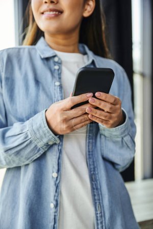 abgeschnittene Ansicht einer lächelnden Geschäftsfrau in blauem Jeanshemd, die mit dem Handy vernetzt ist, während sie im modernen Büro vor verschwommenem Hintergrund steht, junge Unternehmerin, erfolgreiche Jugend